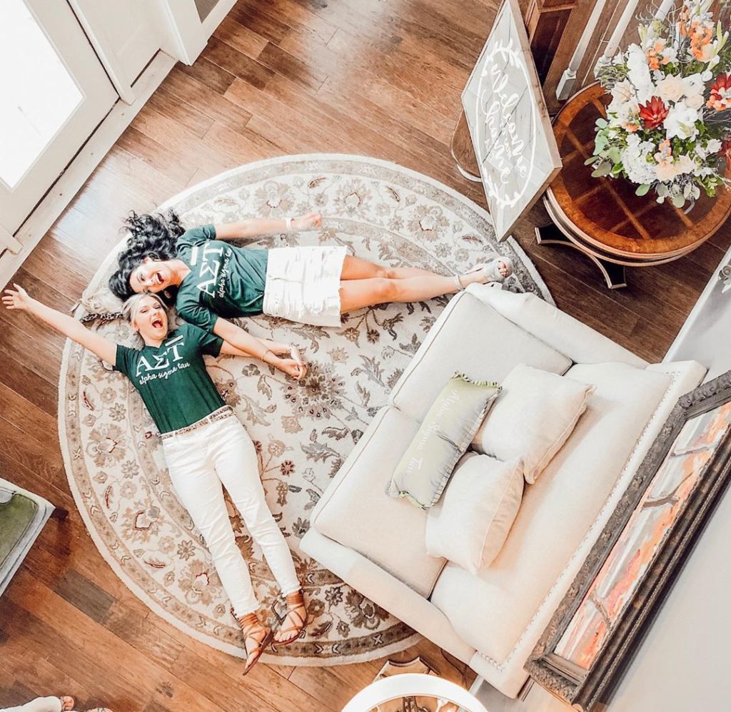 Two women in green Alpha Sigma Tau shirts and white pants and skirt lay on the living room floor, hold hands, and smile at the camera above.